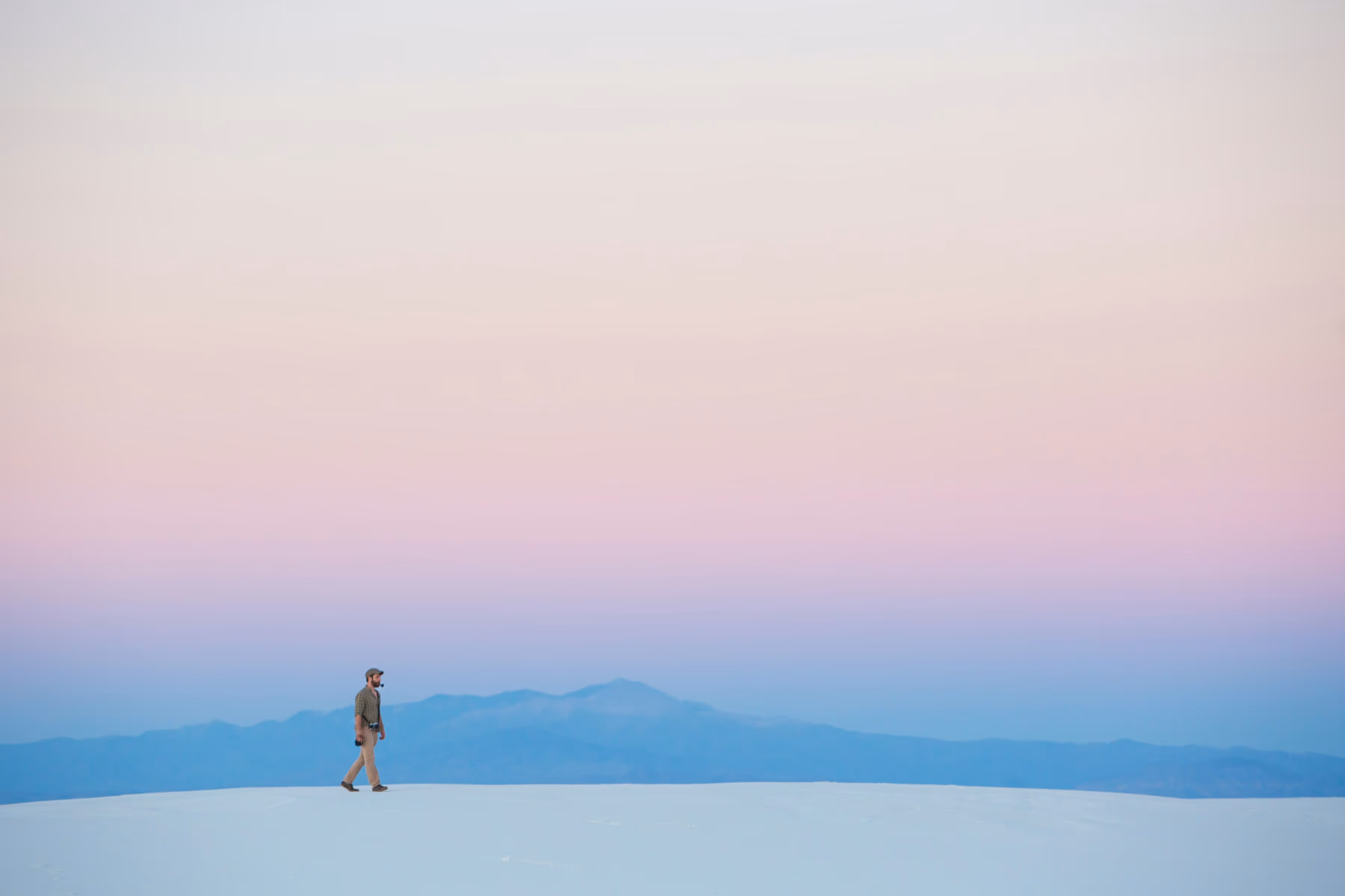 Mann, der bei Sonnenuntergang über weiße Sanddünen geht, dahinter blaue Berge und ein rosa Himmel.